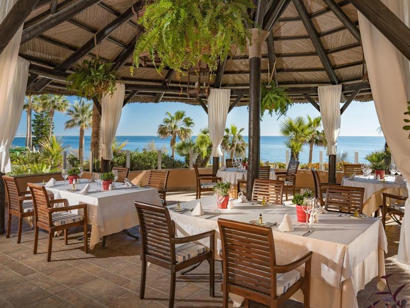 Offenes Restaurant mit Holzstühlen, weißen Tischtüchern und Meerblick bei Sonnenschein.