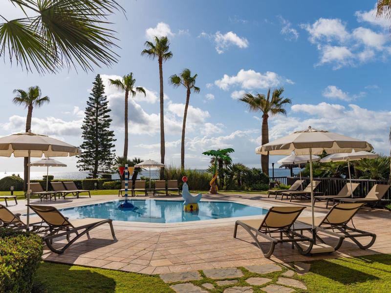 Schwimmbad mit Liegestühlen, Sonnenschirmen und Palmen unter blauem Himmel mit weißen Wolken.