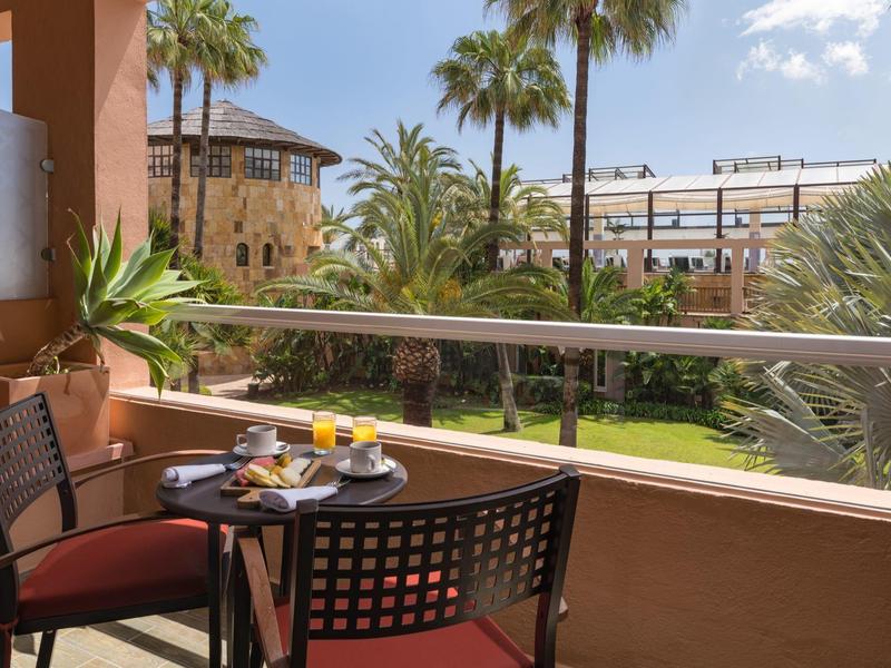 Balkon mit Tisch und Stühlen, Frühstücksgeschirr, Palmen und blauer Himmel im Hintergrund.