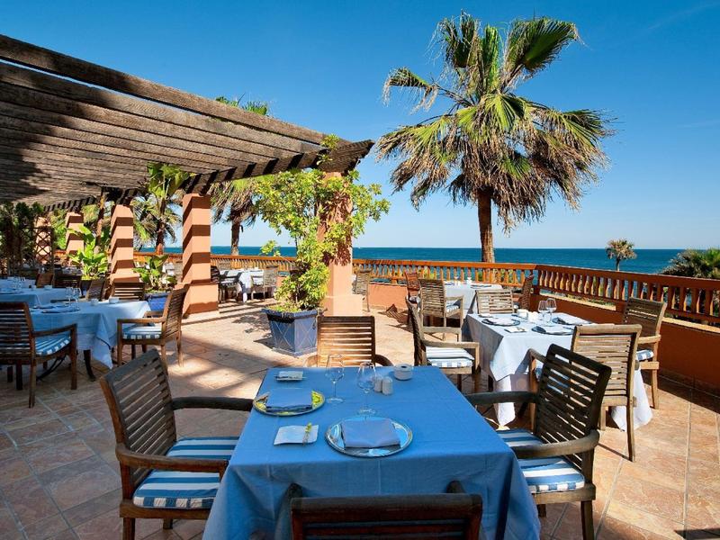 Terrassenrestaurant mit Tischen, Stühlen und Blick auf das Meer bei sonnigem Wetter.