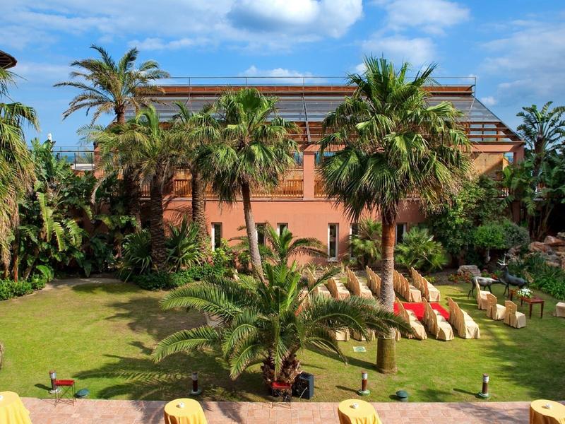 Garten mit Palmen und Liegestühlen vor einem orangefarbenen Hotelgebäude unter blauem Himmel.