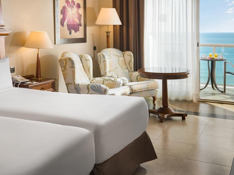 Hotelzimmer mit zwei Einzelbetten, Sesseln, Tisch, Balkon und Meerblick bei Tageslicht.