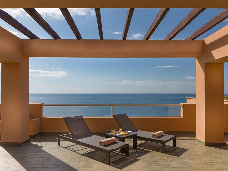 Terrassenhotel mit zwei Liegen und Blauem Meer im Hintergrund unter Pergola bei sonnigem Himmel.