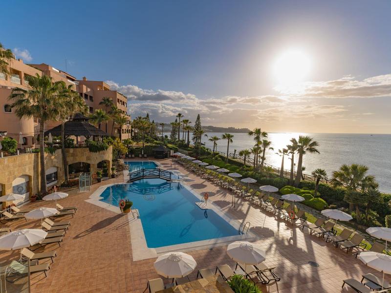 Sonniger Poolbereich mit Liegestühlen, Palmen und Meerblick bei hellem Himmel am späten Nachmittag.