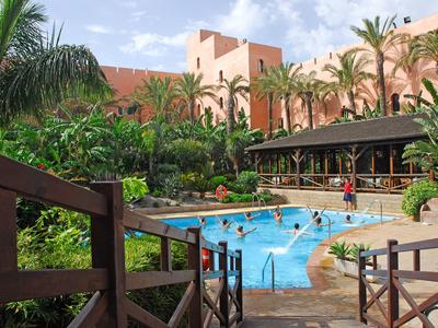 Ein tropischer Pool mit Palmen und roten Gebäuden im Hintergrund unter blauem Himmel.