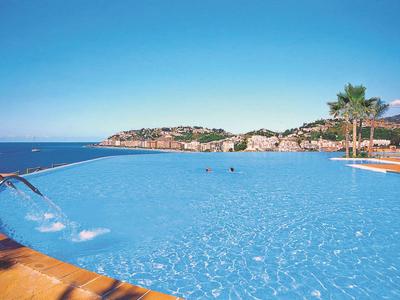 Großer blauer Pool mit Wasserdüsen, Palmen und Felsen im Hintergrund unter klarem Himmel.
