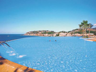 Großer Pool mit Wasserfall, Palmen und Meerblick bei sonnigem Wetter.