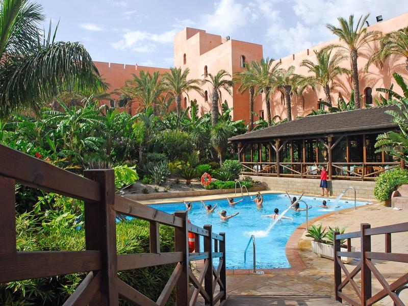 Ein tropischer Pool mit Palmen und roten Gebäuden im Hintergrund unter blauem Himmel.