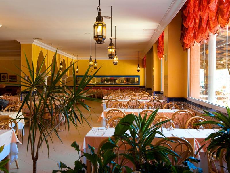 Helles Restaurant mit weißen Tischdecken, orangenen Vorhängen und grünen Pflanzen am Fenster.