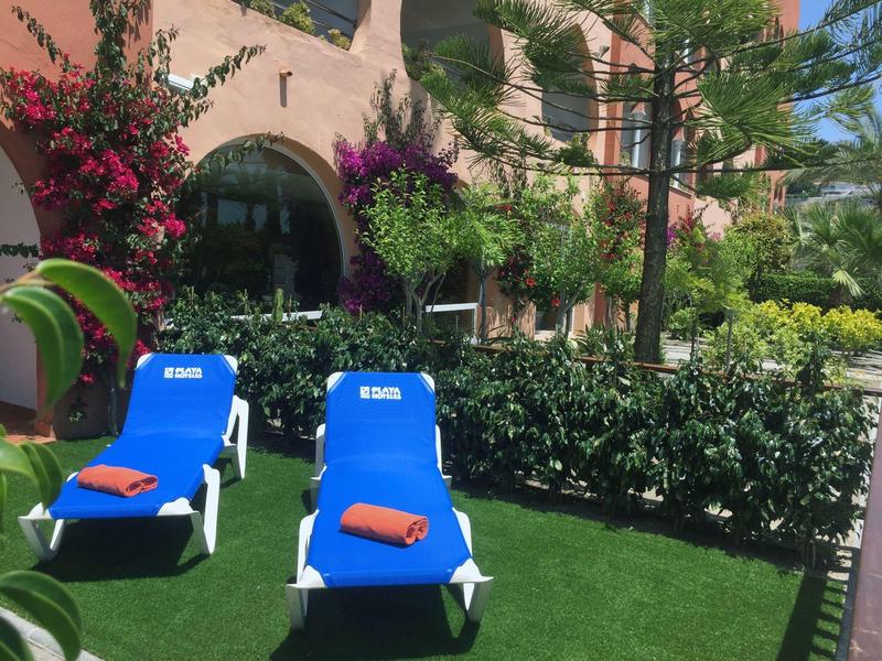 Zwei blaue Liegestühle mit orangenen Handtüchern stehen auf grünem Rasen vor einem rosa Gebäude mit Pflanzen.