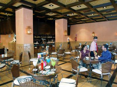 Innenraum eines Restaurants mit Sitzplätzen aus Metall und Glas, Gästen, warmen Wandfarben und Deckenlampen.