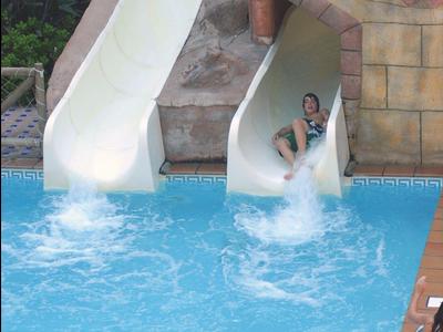 Zwei Kinder rutschen eine weiße Wasserrutsche in einen blauen Pool, umgeben von braunen Fliesen.