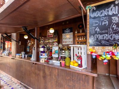 Holzbar mit Cocktailliste, Getränken, bunten Dekorationen und kleinen Ananasfiguren am Tresen.