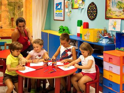 Kinder sitzen am runden roten Tisch und malen, während eine Frau sie beobachtet. Bunte Wände und Möbel.