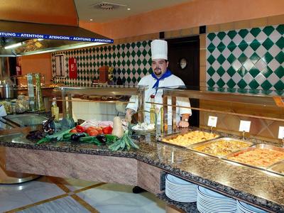 Buffet mit verschiedenen Speisen, ein Koch in weißer Kleidung hinter Theke mit grün-weißen Kacheln.