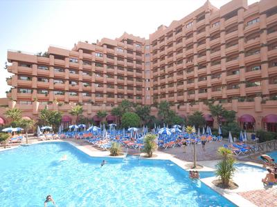 Hotel mit mehrstöckigem Gebäude und großem Außenpool mit Liegestühlen und Sonnenschirmen.