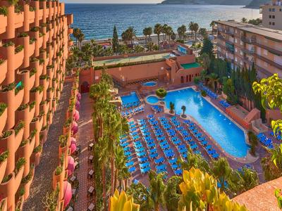 Großes Hotel mit blauem Pool, vielen Liegestühlen und Palmen, Meer im Hintergrund bei Sonnenuntergang.