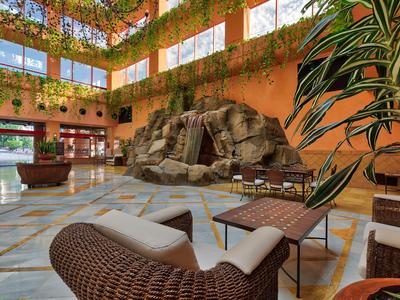 Hotel-Lobby mit hohen Decken, viel Tageslicht, großen Pflanzen und gemütlichen Sitzbereichen.