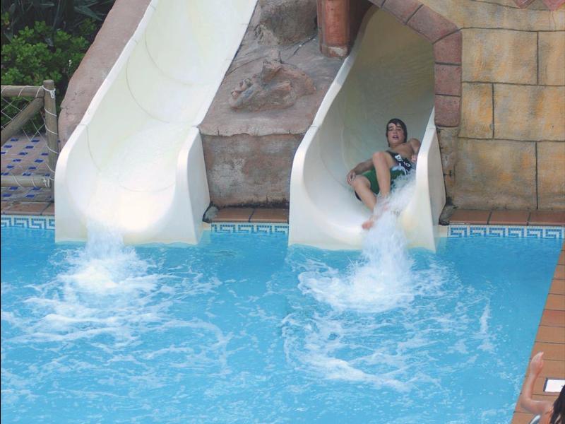 Zwei Kinder rutschen eine weiße Wasserrutsche in einen blauen Pool, umgeben von braunen Fliesen.