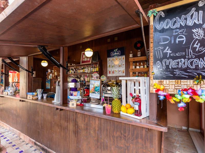 Holzbar mit Cocktailliste, Getränken, bunten Dekorationen und kleinen Ananasfiguren am Tresen.