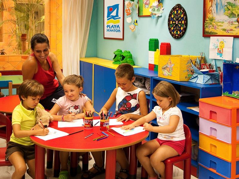 Kinder sitzen am runden roten Tisch und malen, während eine Frau sie beobachtet. Bunte Wände und Möbel.