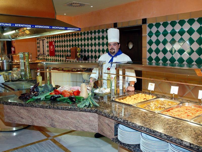 Buffet mit verschiedenen Speisen, ein Koch in weißer Kleidung hinter Theke mit grün-weißen Kacheln.