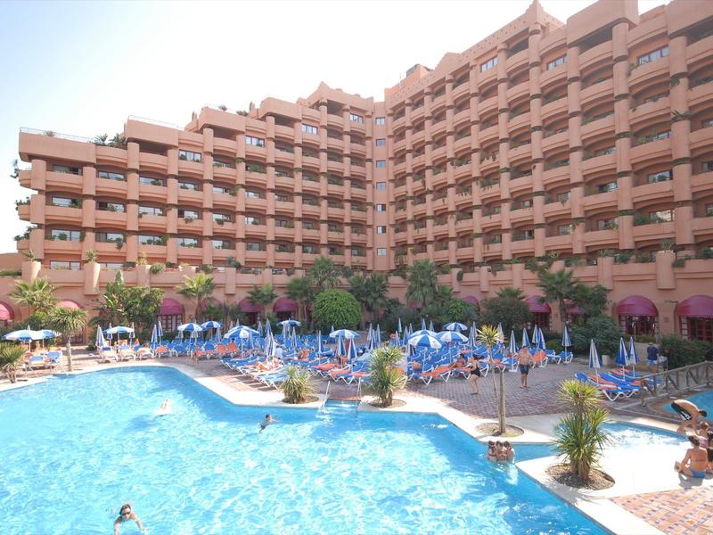Großes Hotelgebäude mit vielen Balkonen, davor blauer Pool und zahlreiche blaue Sonnenliegen.