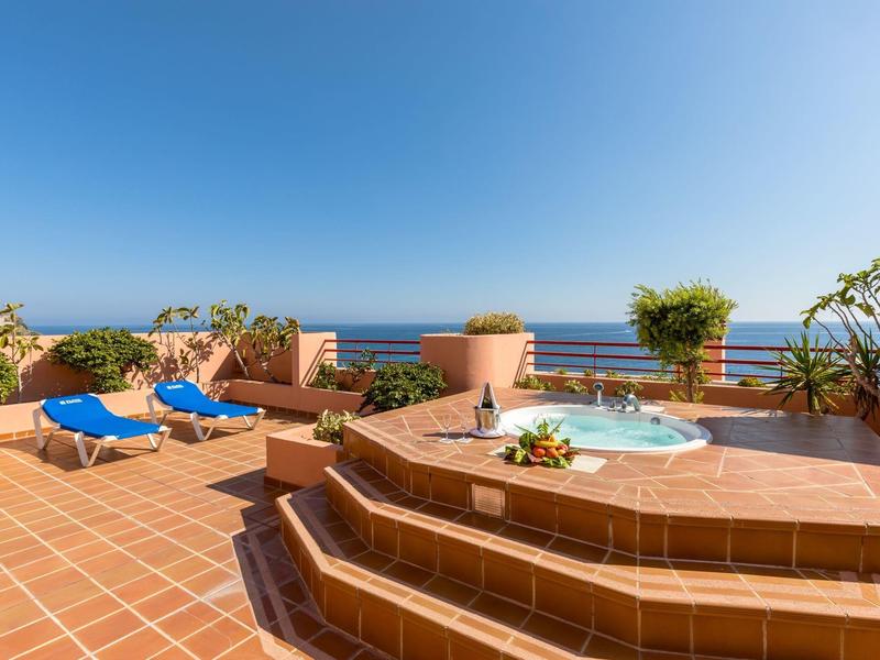 Terrassenbereich mit Jacuzzi, blauer Liege und Blick auf das blaue Meer unter klarem Himmel.