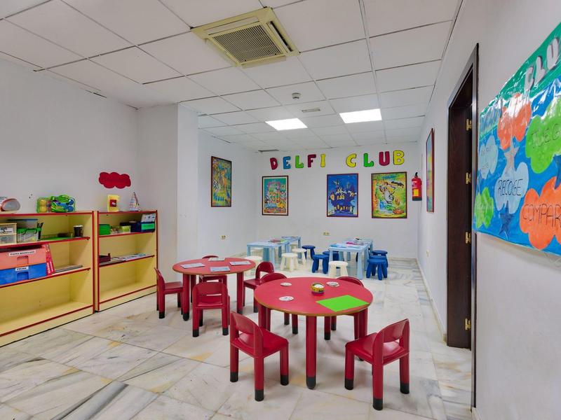 Kinderzimmer mit bunten Tischen und Stühlen, Regal mit Spielzeug und dekorativen Wandbildern.