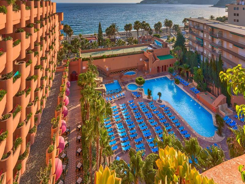 Großes Hotel mit blauem Pool, vielen Liegestühlen und Palmen, Meer im Hintergrund bei Sonnenuntergang.