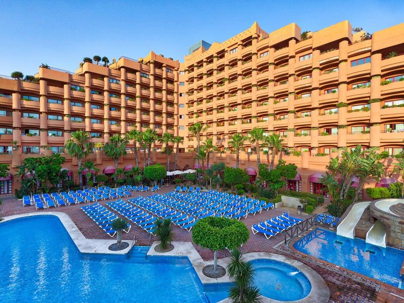 Großes Hotel mit mehreren Stockwerken, vielen blauen Liegen und einem großen Außenpool.