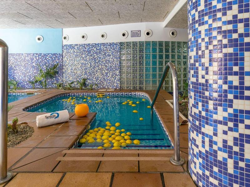 Hallenbad mit Mosaikfliesen, Treppe, Früchten im Wasser und Handtuch am Beckenrand.