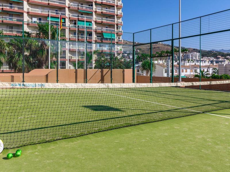 Leerer Tennisplatz mit grünem Kunstrasen, Netz, Tennisschlägern und Bällen neben einem Hochhaus.