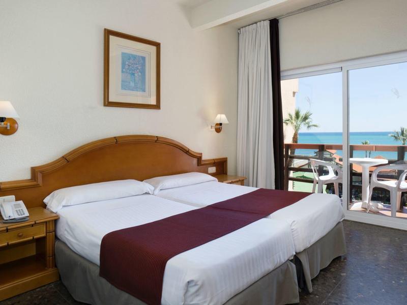 Hell eingerichtetes Hotelzimmer mit Doppelbett, Holzmöbeln und Blick auf Terrasse und Meer.