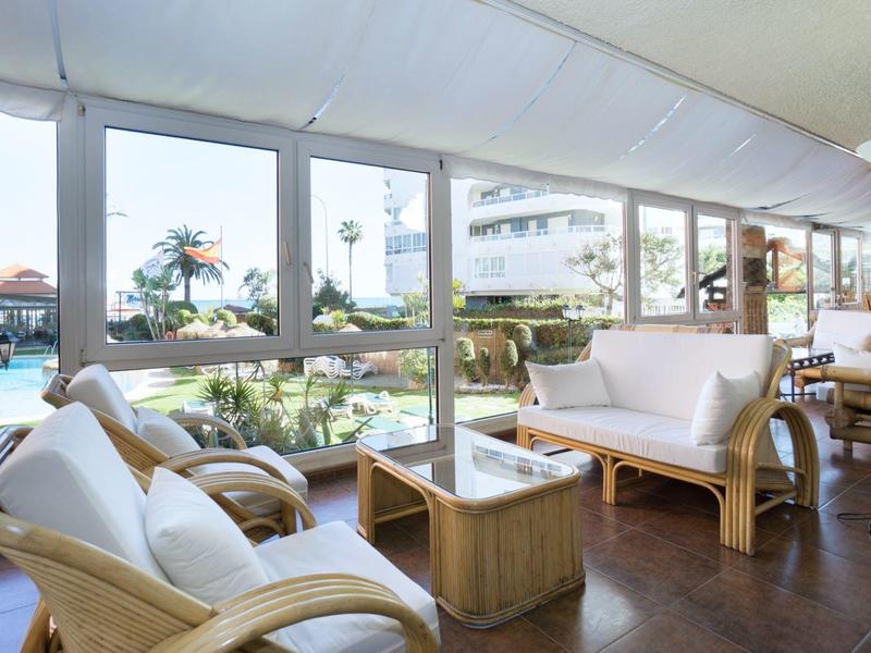 Wohnzimmer mit großen Fenstern, hellen Rattanmöbeln und Blick auf einen Pool und Garten.