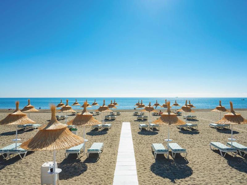 Breiter Sandstrand mit Liegen und Sonnenschirmen, blauem Himmel und ruhigem Meer im Hintergrund.