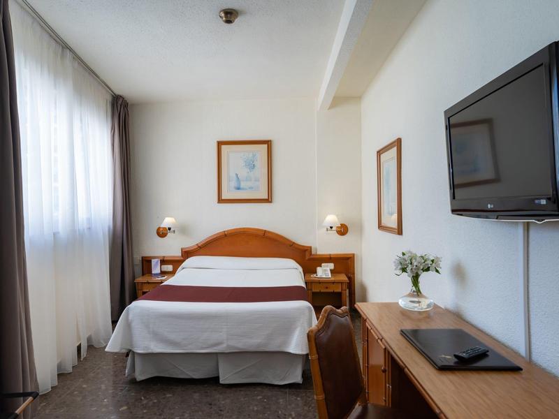 Kleines Hotelzimmer mit Doppelbett, Holzmöbeln, großem Fenster und Wandfernseher.