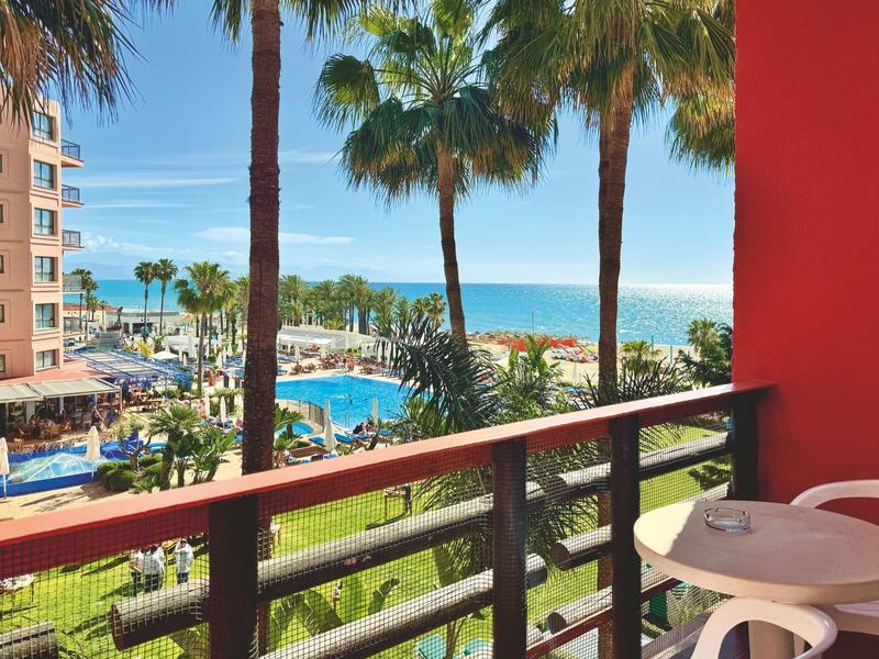 Blick von Balkon auf Palmen, Pool, Gebäude und am Horizont das blaue Meer unter klarem Himmel.