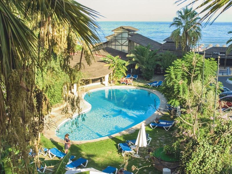 Ein runder blauer Pool umgeben von grünen Palmen und Liegestühlen, im Hintergrund das Meer.