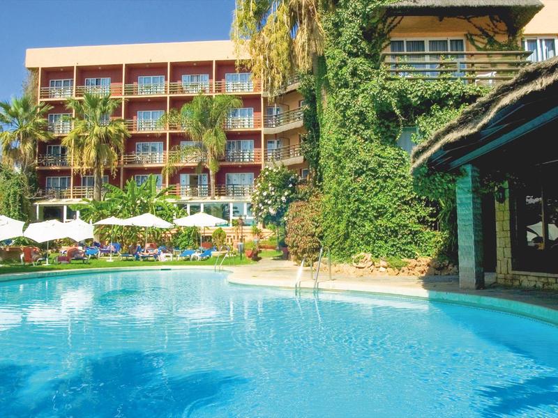 Großer blauer Pool vor mehrstöckigem Hotel mit Palmen und Sonnenschirmen bei sonnigem Wetter.