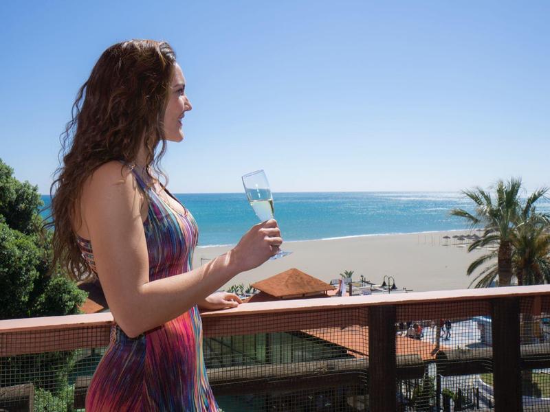 Frau mit langem Haar und sommerlichem Kleid blickt mit Glas in der Hand aufs Meer bei klarem Himmel.