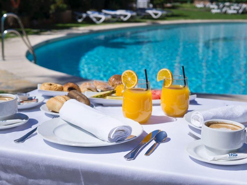 Tisch mit Frühstück, Orangensaft, Kaffee vor blauem Pool und Sonnenliegen im Hintergrund.