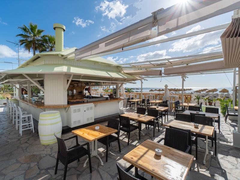 Offene Strandbar mit Holztischen, schwarzen Stühlen und Palmen unter blauem Himmel.