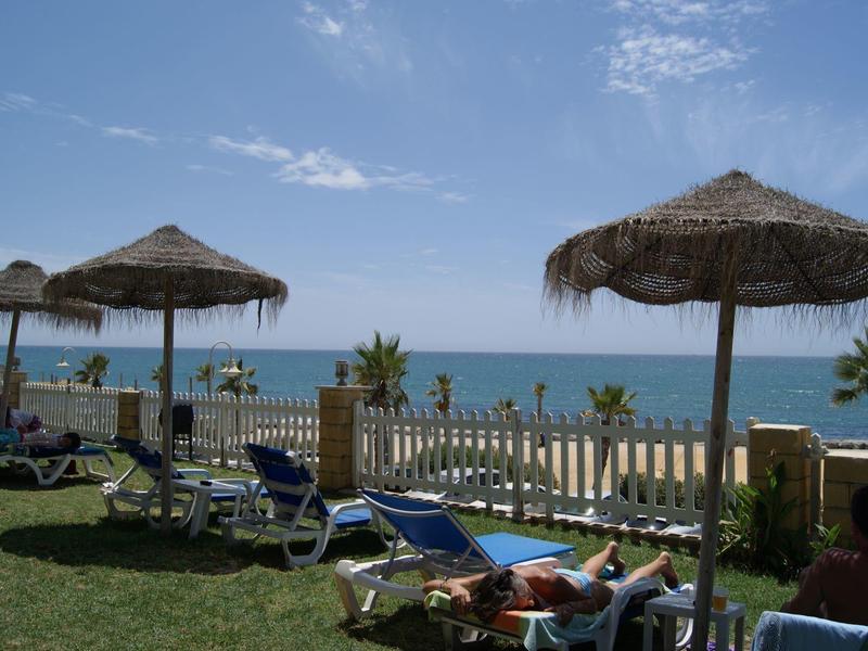 Strand mit Liegestühlen, Strohm Sonnenschirmen, grünem Gras und blauem Meer im Hintergrund.