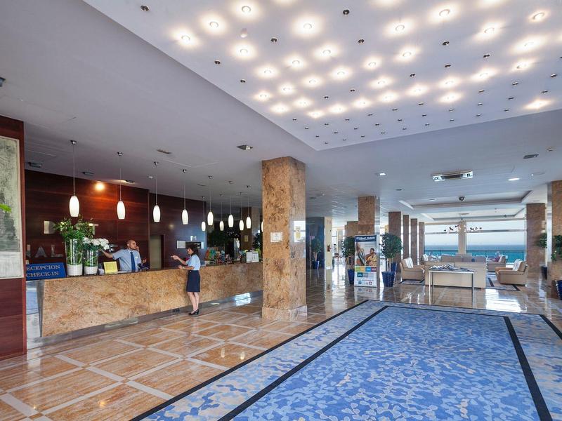 Großer Hotellobby mit blauer Teppichfläche, Rezeption, Sitzbereich und heller Deckenbeleuchtung.