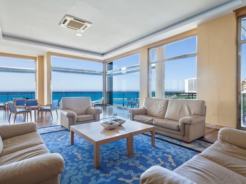 Helles Wohnzimmer mit großen Fenstern, beigen Sofas, blauem Teppich und Meerblick.