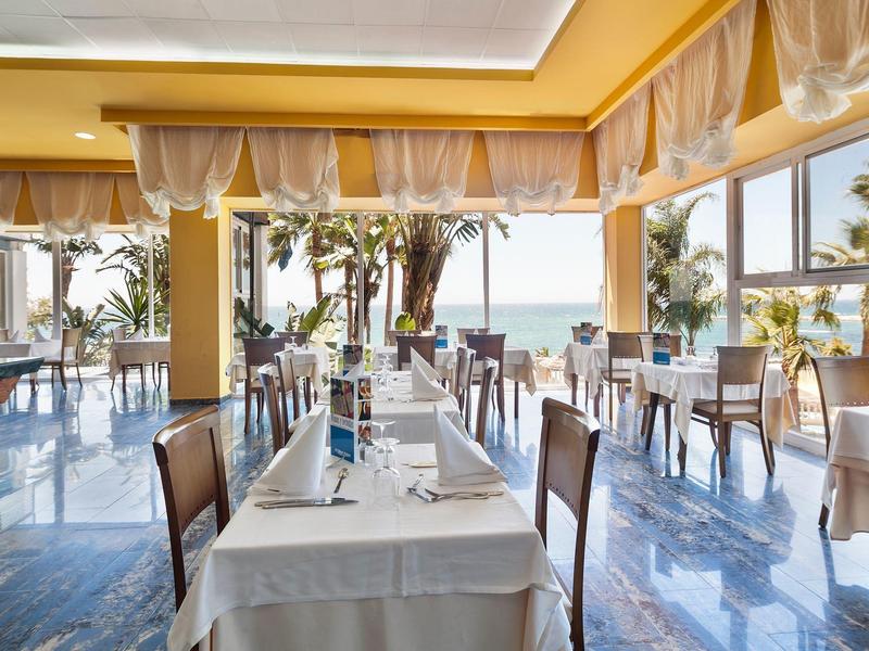 Restaurant mit Meerblick, hellen Wänden, vielen Fenstern, weißen Tischtüchern und dunklen Stühlen.