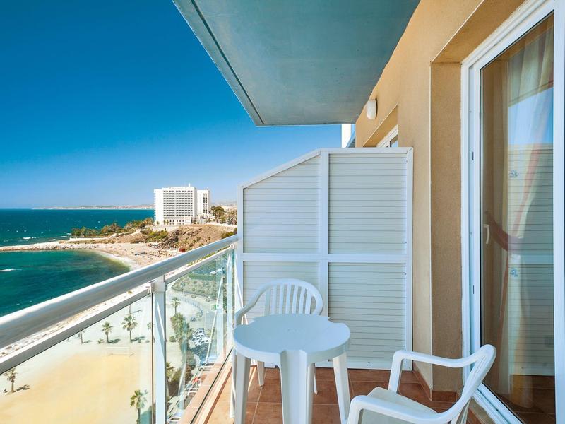 Balkon mit weißem Tisch und Stühlen, Blick auf Strand, Meer und Gebäude unter blauem Himmel.