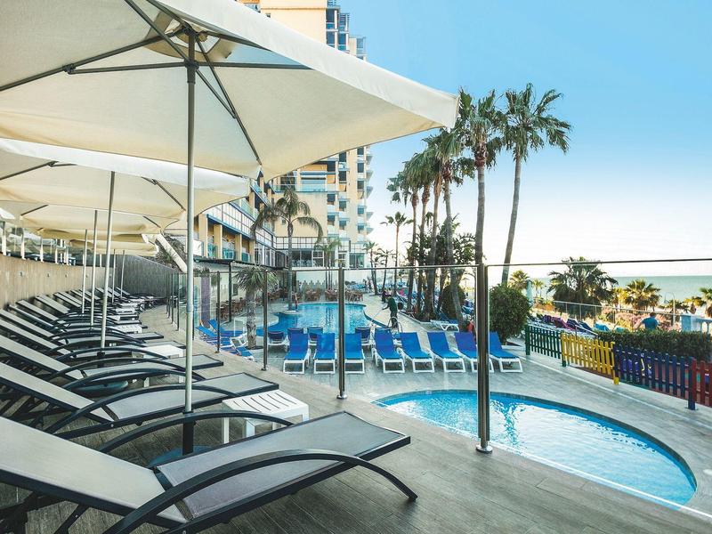 Strandhotel mit Liegestühlen, Sonnenschirmen, Palmen und kleinem Pool am Meer bei klarem Himmel.