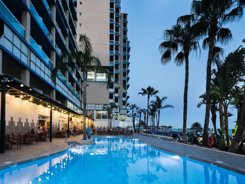 Großer beleuchteter Pool neben modernem Hochhaus mit Palmen und blauer Abenddämmerung.